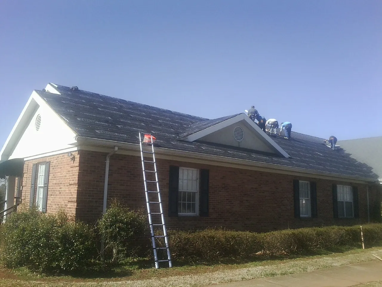 Crew tearing shingles off a brick ranch roof with a ladder set — Metal Roof Repair work by our crew in Lansing